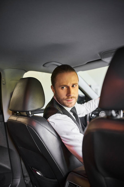 atractivo hombre barbudo en chaleco negro mirando hacia atrás mientras está detrás del volante, concepto de negocio - Foto, imagen