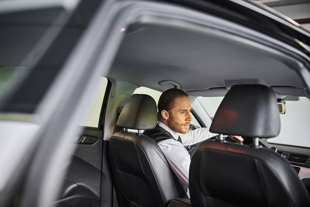 όμορφος κοκκινομάλλης επιχειρηματίας με μαύρο γιλέκο κάθεται πίσω από το τιμόνι και κοιτάζει αλλού - Φωτογραφία, εικόνα