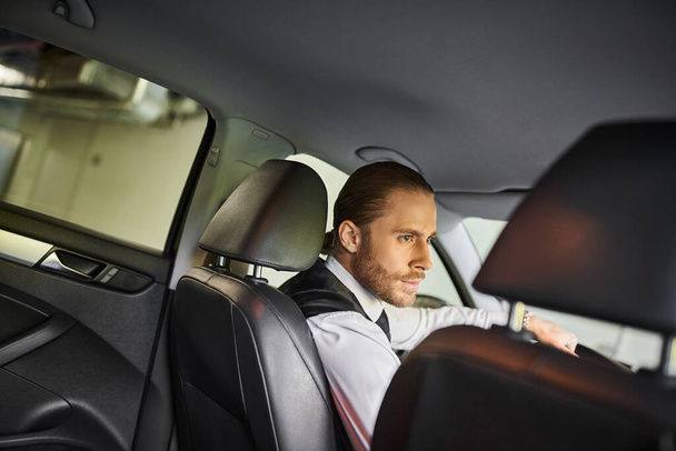 atrakcyjny brodaty mężczyzna z kucykiem w czarnej kamizelce smart za kierownicą, koncepcja biznesu - Zdjęcie, obraz