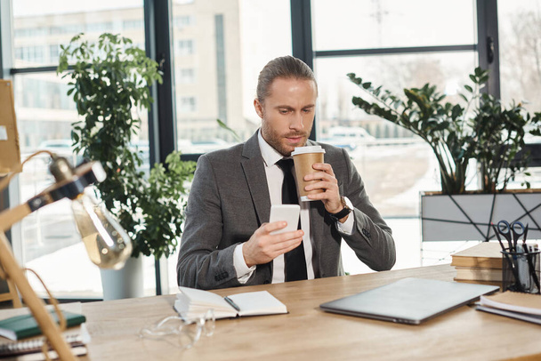 trendy businessman in grey suit drinking coffee to go and chatting on smartphone in office - Photo, Image