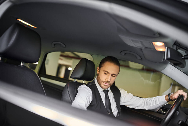 hombre elegante de buen aspecto en chaleco negro detrás del volante en el estacionamiento, concepto de negocio - Foto, imagen