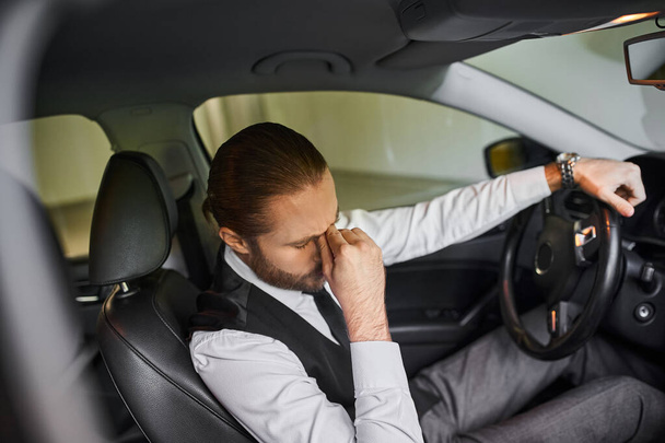 уставший бородатый бизнесмен с хвостиком в черном жилете сидит за рулем своей машины - Фото, изображение