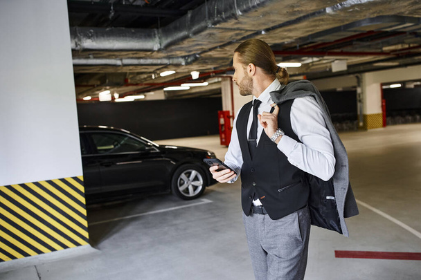 homem barbudo bonito em colete preto segurando telefone celular e olhando para o seu carro, conceito de negócio - Foto, Imagem