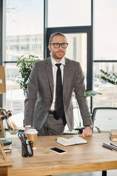 σοβαρός και κομψός επιχειρηματίας με γυαλιά ηλίου που στέκεται στο γραφείο του και κοιτάζει αλλού - Φωτογραφία, εικόνα