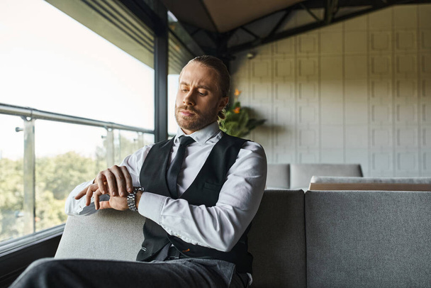 goed uitziende professional in zwart vest zitten en kijken naar zijn polshorloge, business concept - Foto, afbeelding