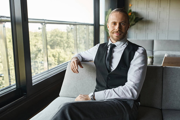 radosny atrakcyjny biznesmen w eleganckim stroju siedzi na kanapie i uśmiecha się lekko do kamery - Zdjęcie, obraz