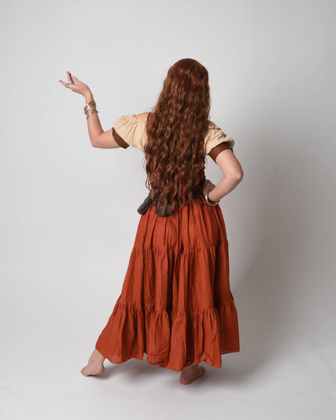 Ολο το μήκος πορτρέτο της όμορφης κοκκινομάλλας γυναίκας που φοράει μια μεσαιωνική παρθένα, κοστούμι μάντη. Στέκεται πίσω θέα, περπατώντας μακριά. απομονωμένο σε φόντο στούντιο. - Φωτογραφία, εικόνα