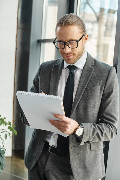 χαμογελαστός επιχειρηματίας με γκρι κοστούμι και γυαλιά που γράφει στο σημειωματάριο ενώ στέκεται στο γραφείο - Φωτογραφία, εικόνα