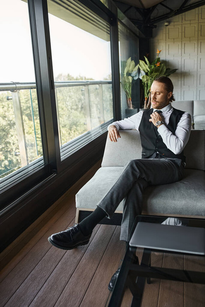 homem de negócios elegante de boa aparência com rabo de cavalo em elegante colete preto inteligente sentado no sofá - Foto, Imagem