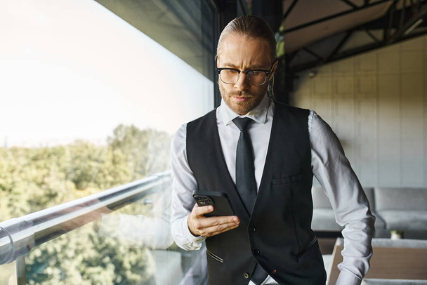 atractivo hombre de negocios con estilo en chaleco negro inteligente mirando el teléfono móvil de pie cerca de la ventana - Foto, Imagen