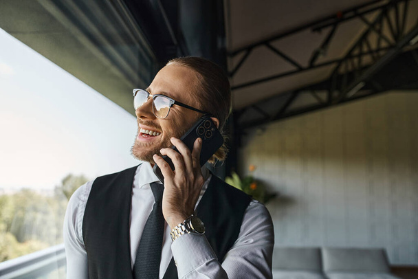 alegre hombre guapo en chaleco negro hablando felizmente por teléfono cerca de la ventana, concepto de negocio - Foto, Imagen
