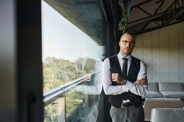 симпатичный бизнесмен в черном жилете и галстуке смотрит в камеру, стоящую рядом с окном - Фото, изображение