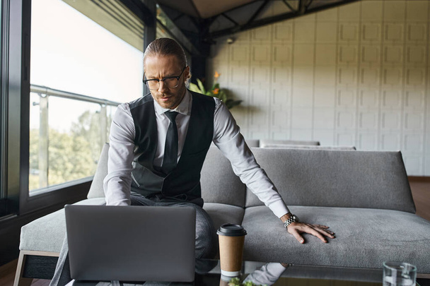 elegante hombre de negocios guapo en chaleco negro con gafas mirando a su computadora portátil, taza de café en la mesa - Foto, Imagen