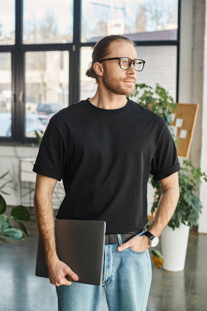 Siyah tişörtlü ve gözlüklü bir şirket yöneticisi dizüstü bilgisayarla ofiste el ele duruyor. - Fotoğraf, Görsel