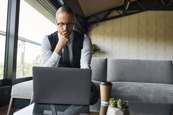 pensive elegante zakenman in zwart vest werken op zijn laptop aandachtig, koffiekop op tafel - Foto, afbeelding