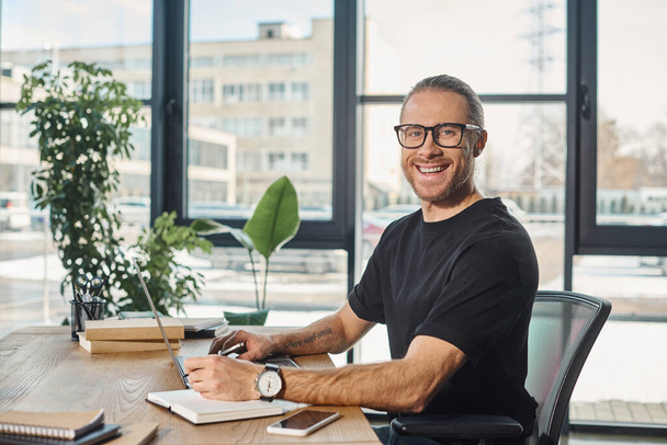 radostný podnikatel v černém tričku a brýlích při pohledu do kamery poblíž notebooku na pracovním stole - Fotografie, Obrázek