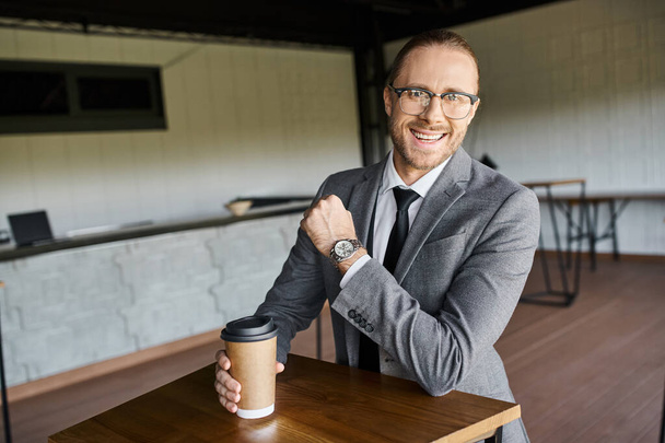 fröhlich gutaussehender Mann im grauen Anzug lächelt in die Kamera und hält Kaffeetasse, Geschäftskonzept - Foto, Bild