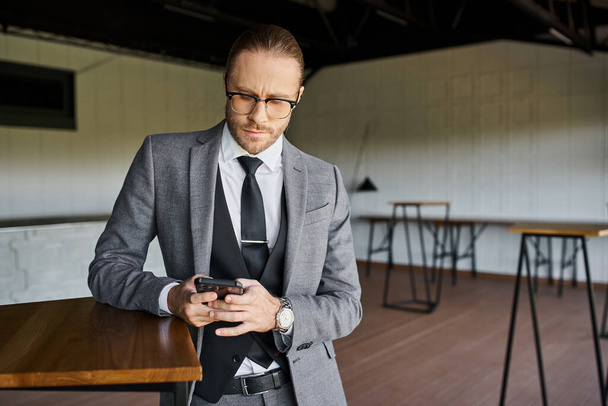 elegante zakenman met bril in slim chique pak die aandachtig naar zijn mobiele telefoon kijkt - Foto, afbeelding