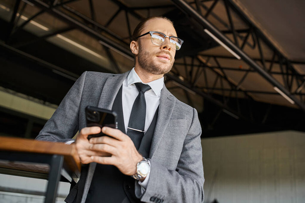 привлекательный элегантный бизнесмен с опрятным стилем с очками и галстуком держа телефон и глядя в сторону - Фото, изображение