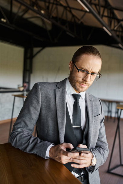 σκεπτικός όμορφος επαγγελματίας με γυαλιά σε γκρι κοστούμι κοιτάζοντας το κινητό του τηλέφωνο, επιχείρηση - Φωτογραφία, εικόνα