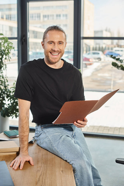 glimlachende zakenman in bril zittend op het bureau met map en lachend naar de camera op kantoor - Foto, afbeelding