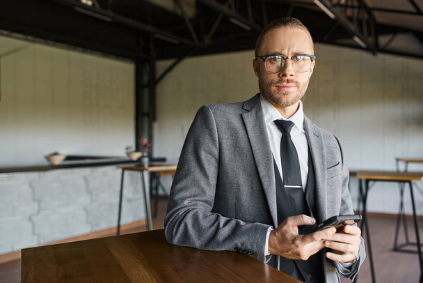 pohledný profesionál s brýlemi v šedém elegantním obleku drží mobilní telefon a dívá se do kamery - Fotografie, Obrázek