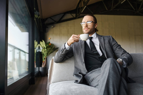 attraktiver Mann mit Brille im grauen Anzug, Tee trinkend auf dem Sofa und wegschauend, Geschäftskonzept - Foto, Bild