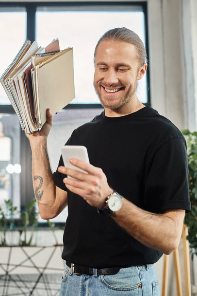 радостный бизнесмен в повседневной одежде, держащий кучу ноутбуков и сообщений на смартфоне в офисе - Фото, изображение