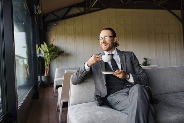 jóképű örömteli profi szürke öltönyben ül a kanapén és teát iszik, üzleti koncepció - Fotó, kép