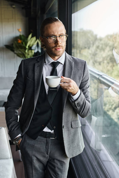 pionowe ujęcie atrakcyjnego eleganckiego biznesmena w szarym garniturze pijącego herbatę z ręką w kieszeni - Zdjęcie, obraz