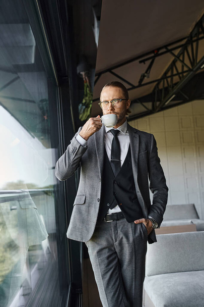 homem de negócios bonito com óculos em terno inteligente bebendo chá e posando com a mão no bolso - Foto, Imagem