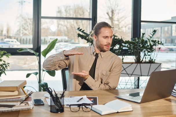 стильный бизнесмен жестикулируя рядом с ноутбуком во время видеозвонка на ноутбуке на рабочем месте в офисе - Фото, изображение