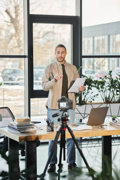 бизнесмен в повседневной одежде стоя с документом рядом с цифровой камерой и ноутбуком, видео маркетинг - Фото, изображение