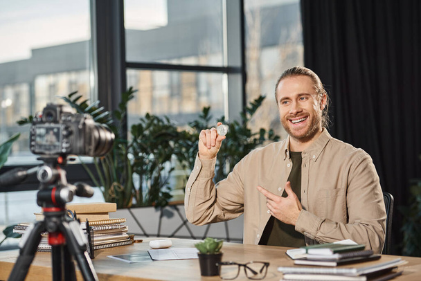 sonriente empresario exitoso mostrando bitcoin durante video blog en el lugar de trabajo en la oficina moderna - Foto, Imagen