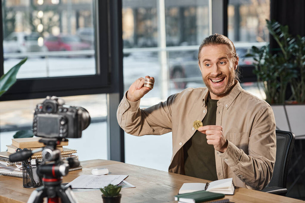 веселый бизнесмен показывает биткоины, сидя на рабочем месте перед цифровой камерой - Фото, изображение