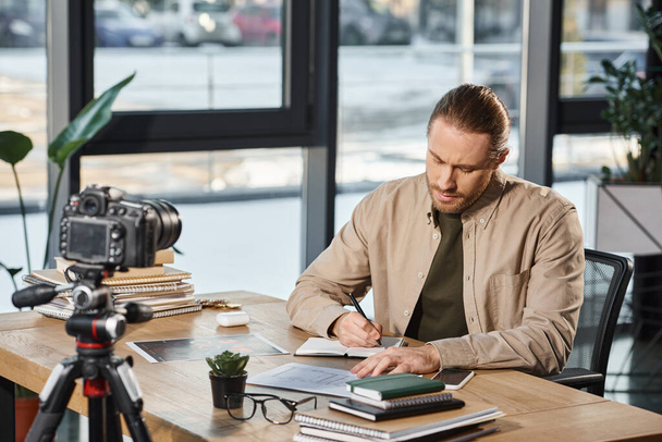 серйозний бізнесмен в повсякденному вбранні, що пише в блокноті на робочому столі перед цифровою камерою - Фото, зображення