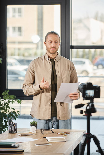 Seriöser Geschäftsmann hält Dokument in der Hand und spricht im modernen Büro vor der Digitalkamera - Foto, Bild