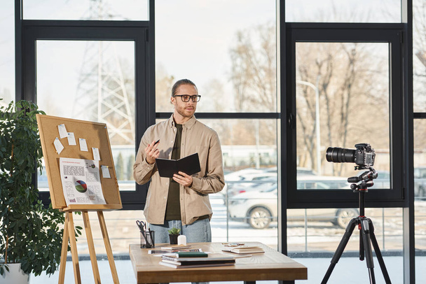 бізнесмен в окулярах тримає блокнот і розмовляє на цифровій камері під час відеоблогу в офісі - Фото, зображення
