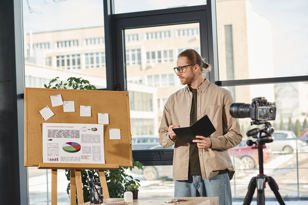менеджер с ноутбуком смотрит на пробковую доску с графиками во время видеопрезентации в современном офисе - Фото, изображение