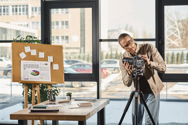 бизнесмен в очках регулирует цифровую камеру возле стола и пробковую доску с графиками в офисе - Фото, изображение