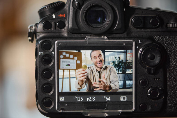 Nahaufnahme einer Digitalkamera in der Nähe eines aufgeregten Unternehmers, der Videoblog am Arbeitsplatz im Büro aufzeichnet - Foto, Bild