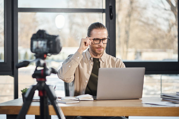 уверенный бизнесмен в очках смотрит на ноутбук перед цифровой камерой в современном офисе - Фото, изображение