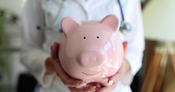 Eine Ärztin mit einem rosafarbenen Sparschwein in der Hand, eine Nahaufnahme. Teure medizinische Verfahren, Investitionen in die Gesundheit - Filmmaterial, Video