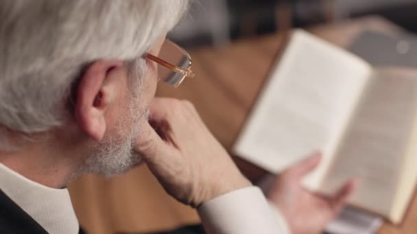 Vue au-dessus de l'épaule des lunettes ajustables pour hommes aux cheveux gris tout en réfléchissant à l'information contenue dans le livre au bureau. Considérer que les dirigeants d'entreprise améliorent la capacité de navigation des relations sociales. - Séquence, vidéo