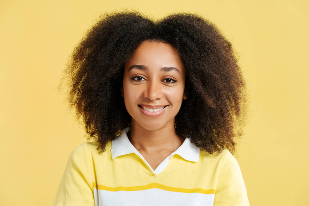Atractiva mujer afroamericana sonriente con el pelo rizado vistiendo camiseta amarilla mirando a la cámara, cara emocional, primer plano aislado en el fondo amarillo. Concepto de belleza natural - Foto, imagen
