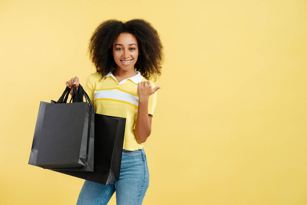 Портрет позитивної афроамериканської жінки з кучерявим волоссям у стильному повсякденному одязі, що тримає чорний пакет для покупок, вказуючи палець на копіювальному просторі, ізольованому на жовтому тлі. концепція Чорної п'ятниці - Фото, зображення