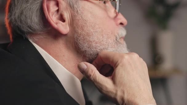 Close-up zijaanzicht van oude man in formeel pak en stropdas streelt zijn baard op kantoor achtergrond. Ervaren ondernemer die psychologisch comfort bereikt tijdens het omgaan met stress op het werk. - Video