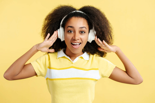 Retrato de una mujer afroamericana emocionada y positiva de pie escuchando música en auriculares mirando a la cámara aislada sobre un fondo amarillo. Concepto tecnológico - Foto, Imagen