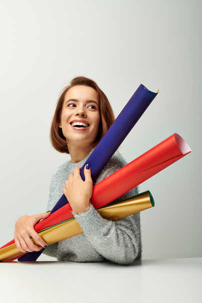 pleased woman with short hair holding colorful wrapping paper on grey backdrop, Christmas concept - Photo, Image