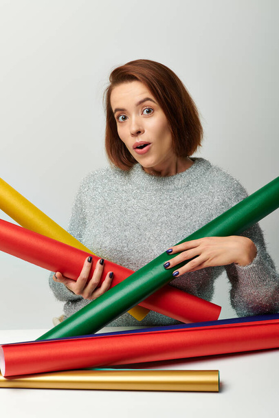 wow emotion, woman in sweater holding colorful wrapping paper on table with grey wall, Christmas - Photo, Image